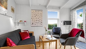 A modern living room with a grey sofa, a wooden coffee table, and a grey armchair. There is a poster of coastal birds on the wall, a small shelf with items, and a window with a view of greenery. - 1 bedroom cozy home in Allinge (Allinge)