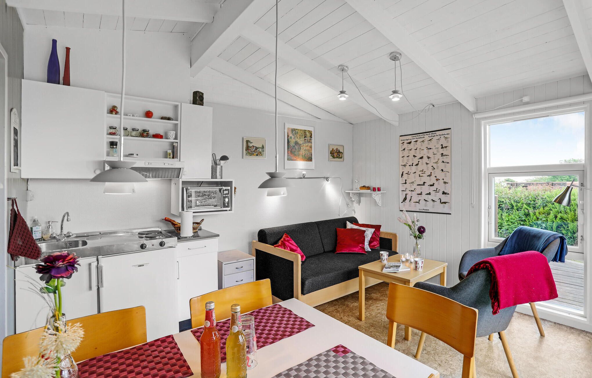 A compact kitchen with modern appliances, a dining area with a table and chairs, a sofa, and a view of greenery through a large window.