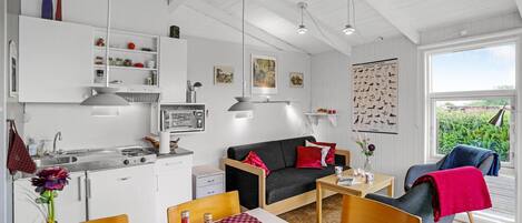 A compact kitchen with modern appliances, a dining area with a table and chairs, a sofa, and a view of greenery through a large window.