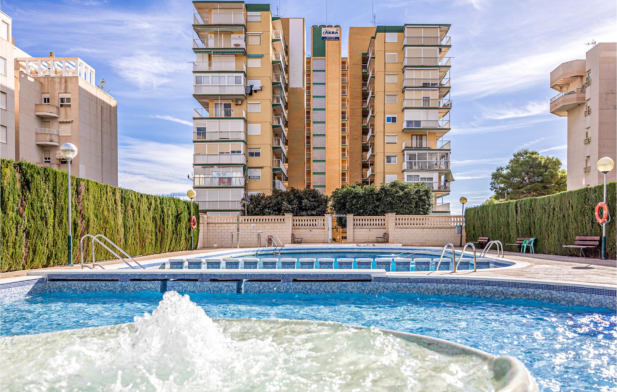 A swimming pool with a fountain, surrounded by a high-rise building with multiple balconies and a hedge.