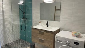 A modern bathroom with a glass shower enclosure, a wooden vanity with a sink, a mirror, and a washing machine. - Elegant, recently renovated apartment (Sainte-Anne)