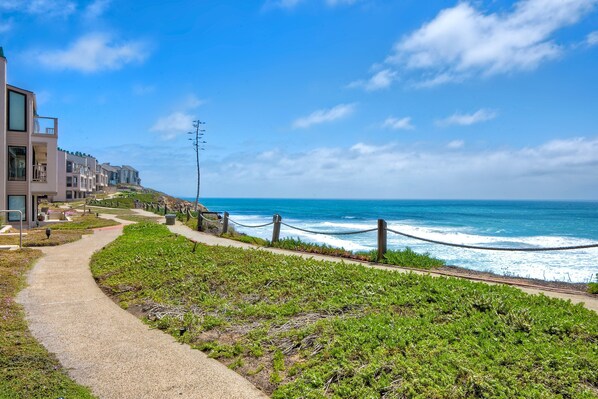 A coastal walkway with a view of the ocean, a modern building, and a grassy area.