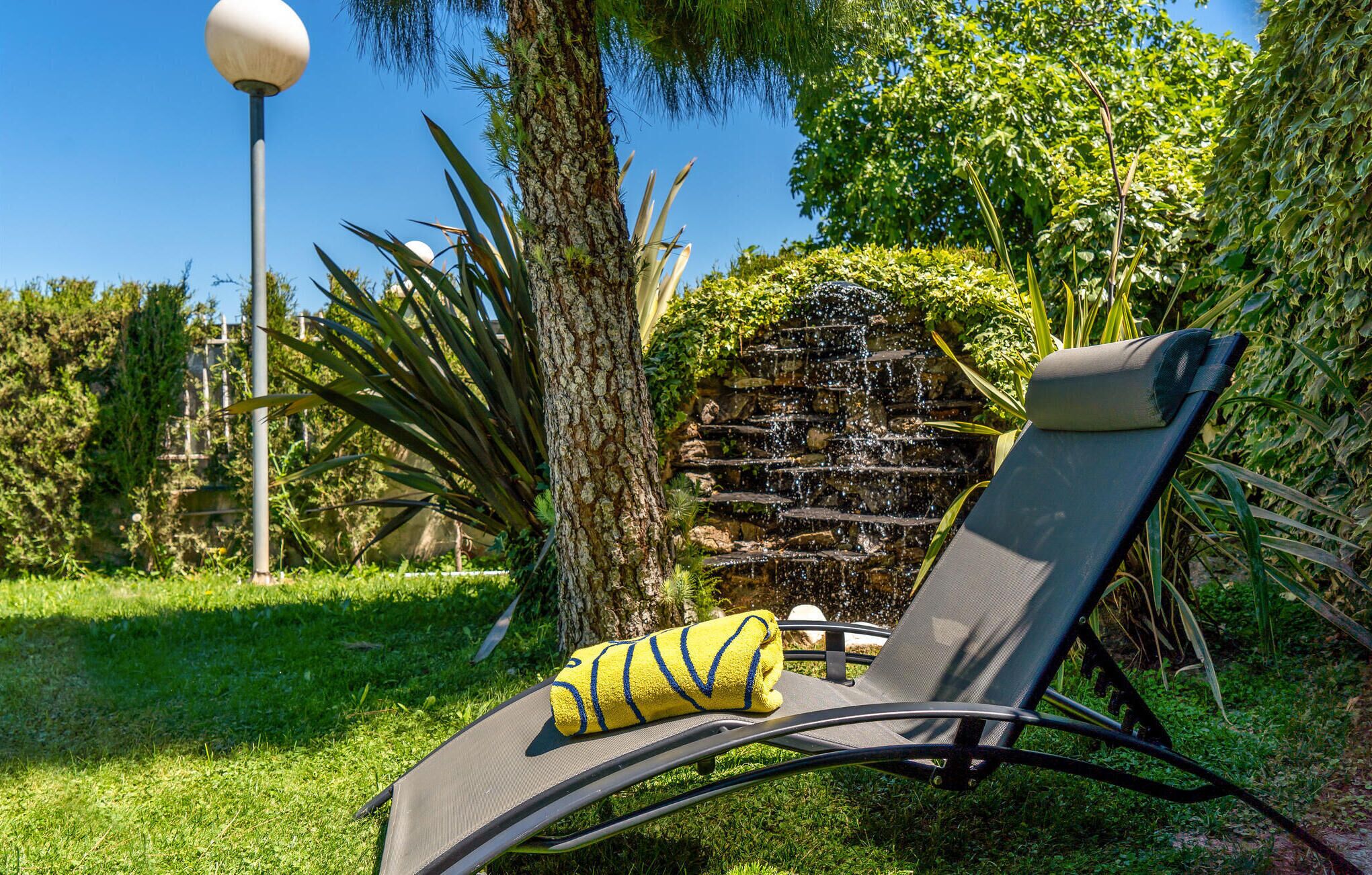 A lounge chair with a towel, a stone water feature, and lush greenery.