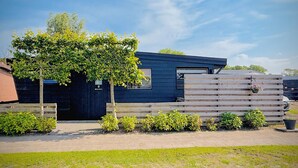 Un bâtiment moderne à la façade sombre, doté d’une terrasse en bois et d’un petit jardin agrémenté de diverses plantes.