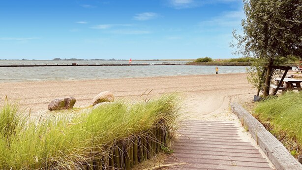 Une promenade en bois mène à une plage de sable, avec une étendue d’eau calme en arrière-plan.