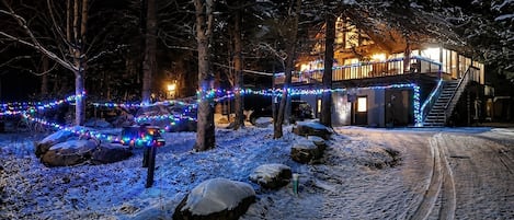 A snow-covered landscape with a house illuminated by string lights, a wooden deck, and a staircase leading to the entrance.