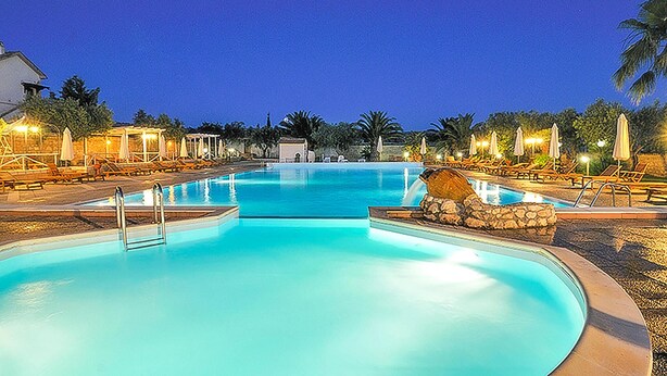 Una piscina amplia y bien iluminada con sillones de descanso y sombrillas, rodeada de palmeras y un muro de piedra.