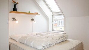 A bedroom with a sloped ceiling, a skylight, a bed with white bedding, two wall-mounted lights, and a shelf with a vase and books. - Cozy apartment in Gudhjem with WiFi (Gudhjem)