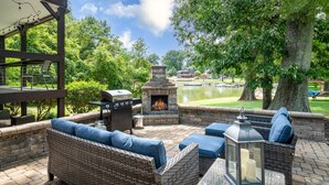 Un patio al aire libre con muebles de mimbre, una chimenea de piedra y vistas a un lago y edificios al fondo.