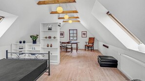 Een ruime slaapkamer op zolder met een hoogslaper, houten balken, een eettafel met stoelen en een groot raam.