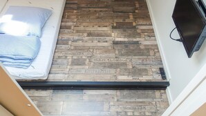 Una habitación estrecha con una cama a la izquierda, un televisor a la derecha y suelo de madera con texto visible.