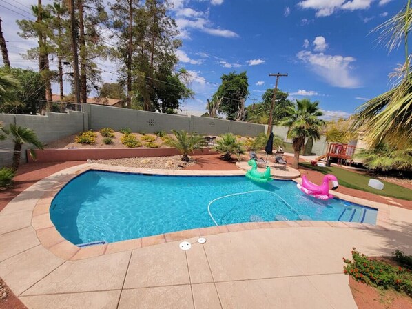 A swimming pool with a curved edge, surrounded by a tiled deck. There are palm trees and a fence in the background. - Spacious 4BR Near Strip with Private Pool, Free Parking (Las Vegas)