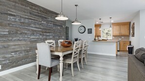 Een eethoek met een houten tafel en stoelen, een keuken op de achtergrond en een woonkamer met een bank.