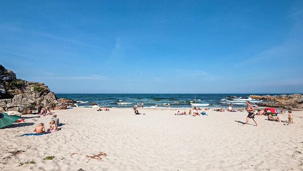 A sandy beach with people sunbathing and swimming, a rocky coastline, and a clear blue sky.