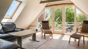 A cozy, well-lit attic room with a sloped ceiling, a black leather sofa, a small table, and two chairs. Large windows offer a view of the outdoors.