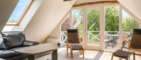A cozy, well-lit attic room with a sloped ceiling, a black leather sofa, a small table, and two chairs. Large windows offer a view of the outdoors.