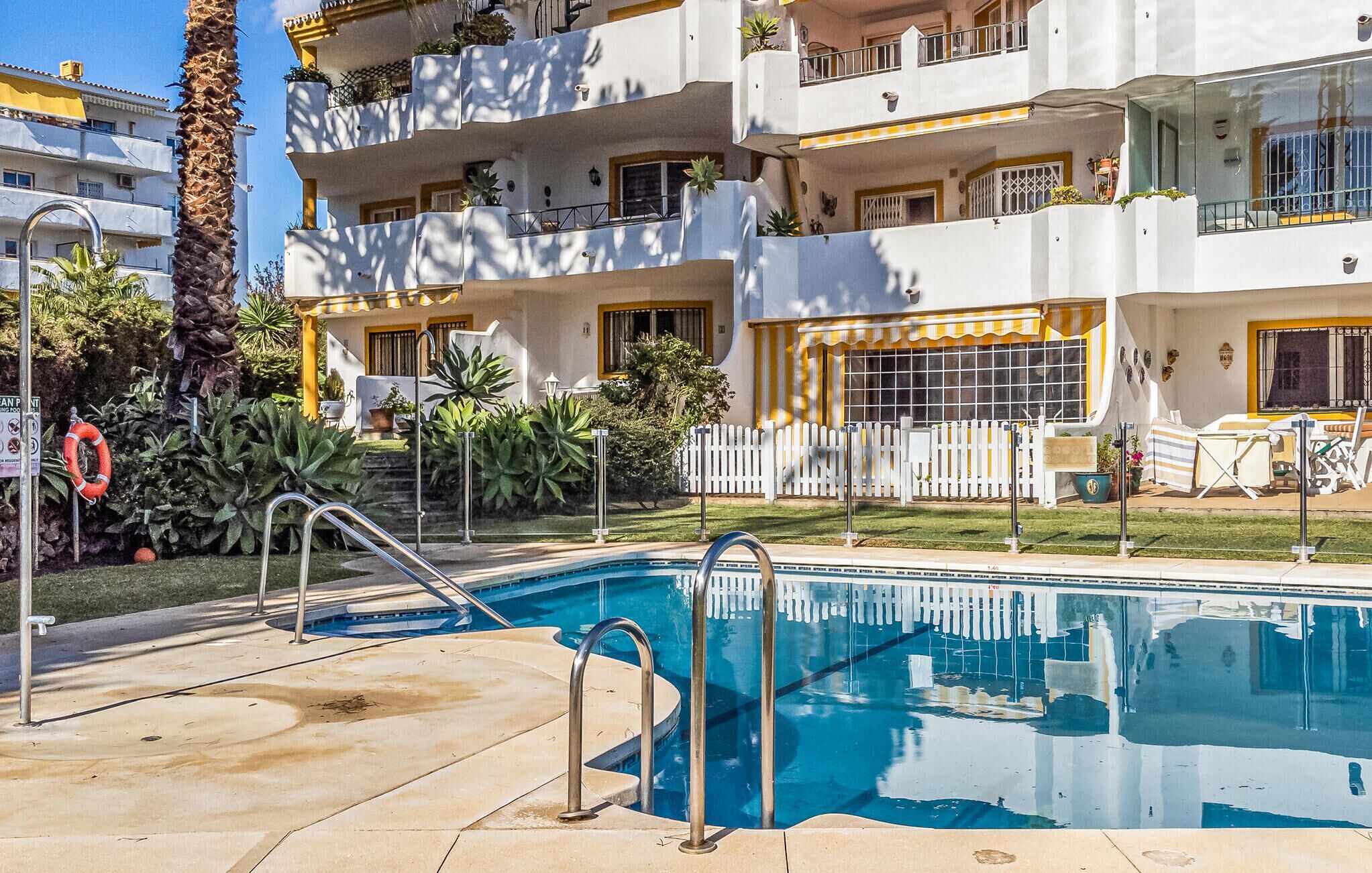 A swimming pool with a white fence, palm trees, and a building with balconies.