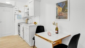 A compact kitchen with white cabinets, a sink, and a dining area with a table and chairs. A framed painting of a street scene hangs on the wall. - Awesome apartment in Nexø with sauna (Nexø)