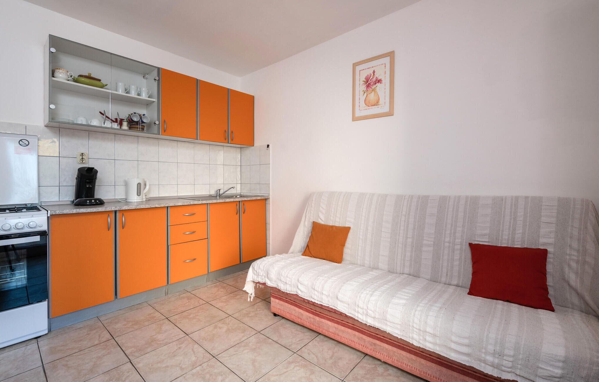 Et lille køkken med orange skabe, en hvid bordplade og en komfur. Der er en sofa med en rød pude og et hvidt tæppe.