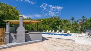 Un espace piscine agrémenté d’une terrasse en pierre, d’un coin barbecue abrité et de chaises longues blanches.