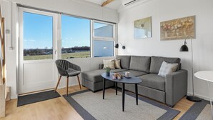 Un salón moderno con un sofá gris, una mesa redonda pequeña y una silla. Amplias ventanas ofrecen vistas al campo.