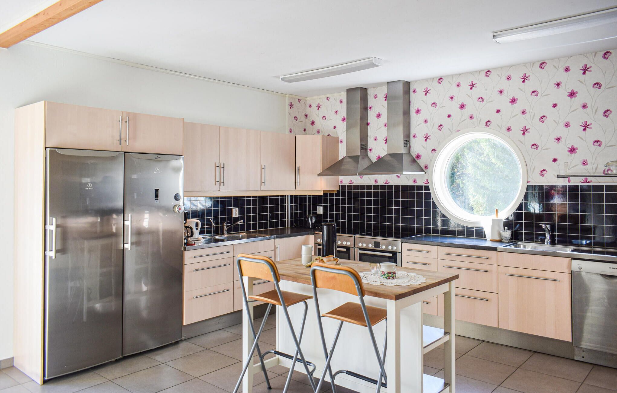 Et moderne køkken med et rustfrit stålkøleskab, træskabe, et rundt vindue og et flisebeklædt backsplash.