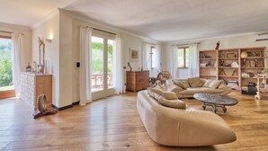 A spacious living room with a large L-shaped sofa, a coffee table, and a wooden floor. There is a bookshelf filled with books and decorative items, and a dining area with a table and chairs visible through the glass doors. - Gorgeous home in Prelà (Prelà)