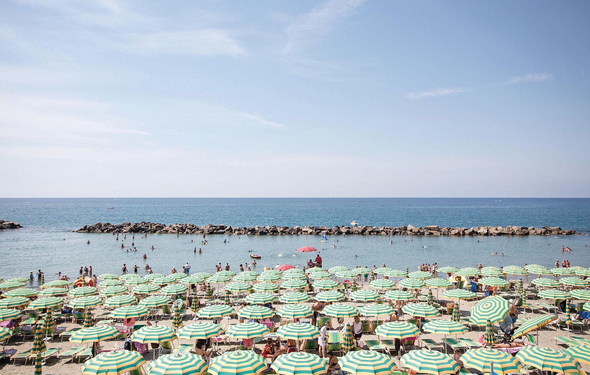 Ein Strand mit zahlreichen gestreiften Sonnenschirmen und Liegestühlen, einem Felssteg und Personen, die schwimmen und entspannen.