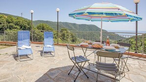 Eine Terrasse mit Esstisch, zwei Sonnenliegen und einem großen, gestreiften Sonnenschirm, der Schatten spendet.