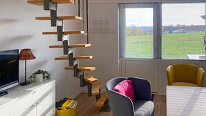 Ein modernes Wohnzimmer mit Treppe, Flachbildfernseher, Esstisch und Blick auf ein grünes Feld durch das Fenster.