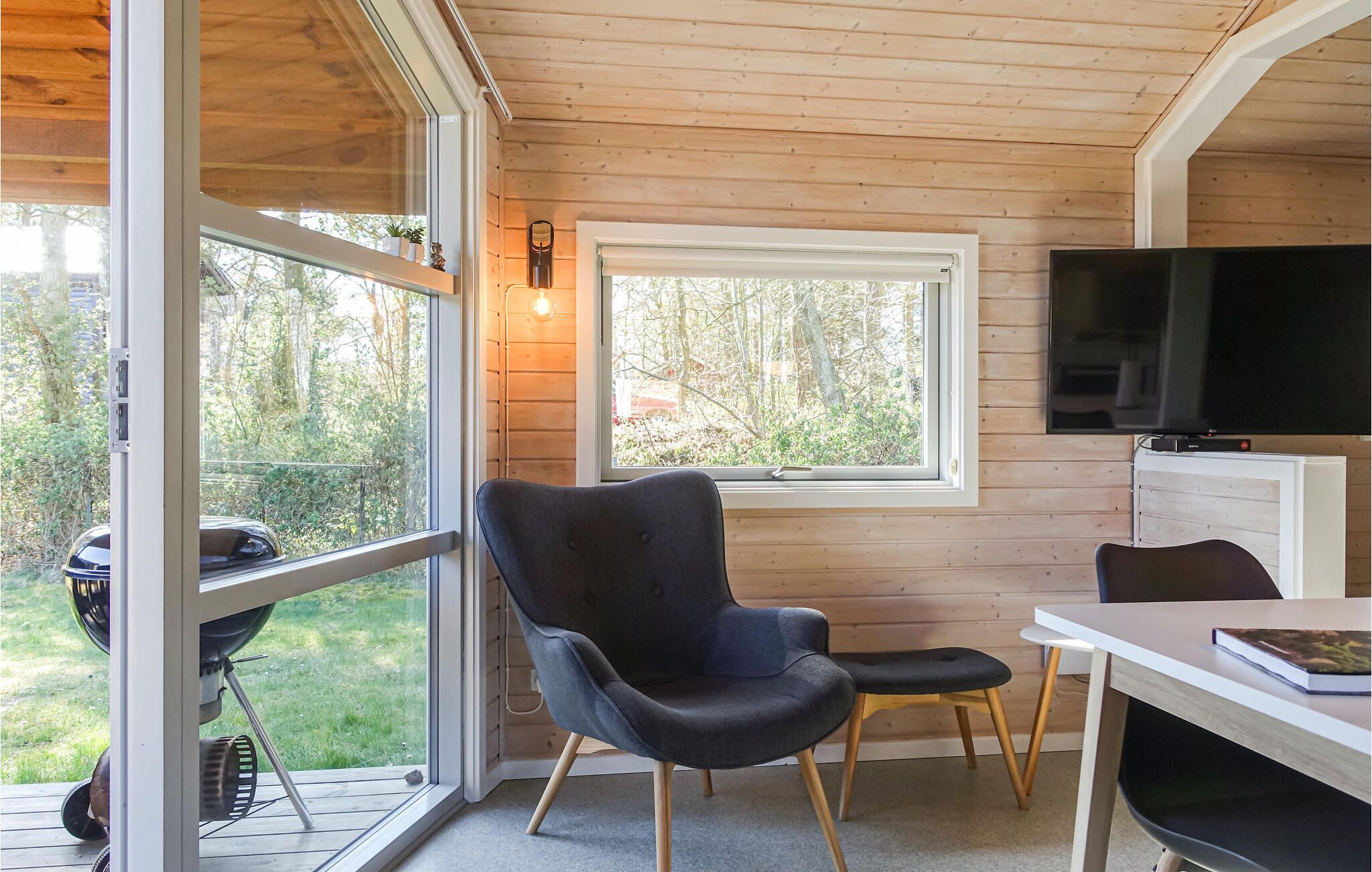 Ein gemütliches Blockhaus-Interieur mit Holzbalkendecke, einem großen Fenster mit Blick auf einen Garten, einem schwarzen Sessel, einem kleinen Tisch und einem an der Wand befestigten Fernseher.