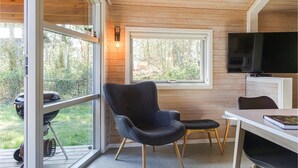 Ein gemütliches Blockhaus-Interieur mit Holzbalkendecke, einem großen Fenster mit Blick auf einen Garten, einem schwarzen Sessel, einem kleinen Tisch und einem an der Wand befestigten Fernseher.
