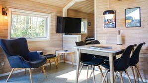 Ein gemütlicher Raum mit Holzvertäfelung, einem an der Wand montierten Fernseher, einem Esstisch mit Stühlen und einem Fenster, das für natürliches Licht sorgt.