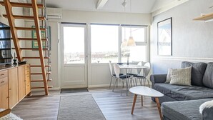Ett modernt vardagsrum med en grå soffa, en trästege, ett matbord med stolar och ett stort fönster som ger utsikt över omgivningen.