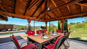An outdoor dining area with a wooden table and red chairs, overlooking a pool and scenic landscape. - Nice home in Ljubescica with sauna (Ljubescica)