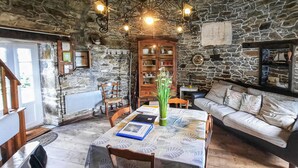 A rustic room with a stone wall, a wooden table with a lace tablecloth, a sofa with patterned cushions, and a chandelier. - 2 bedroom beach front home in Cherrueix (Cherrueix)