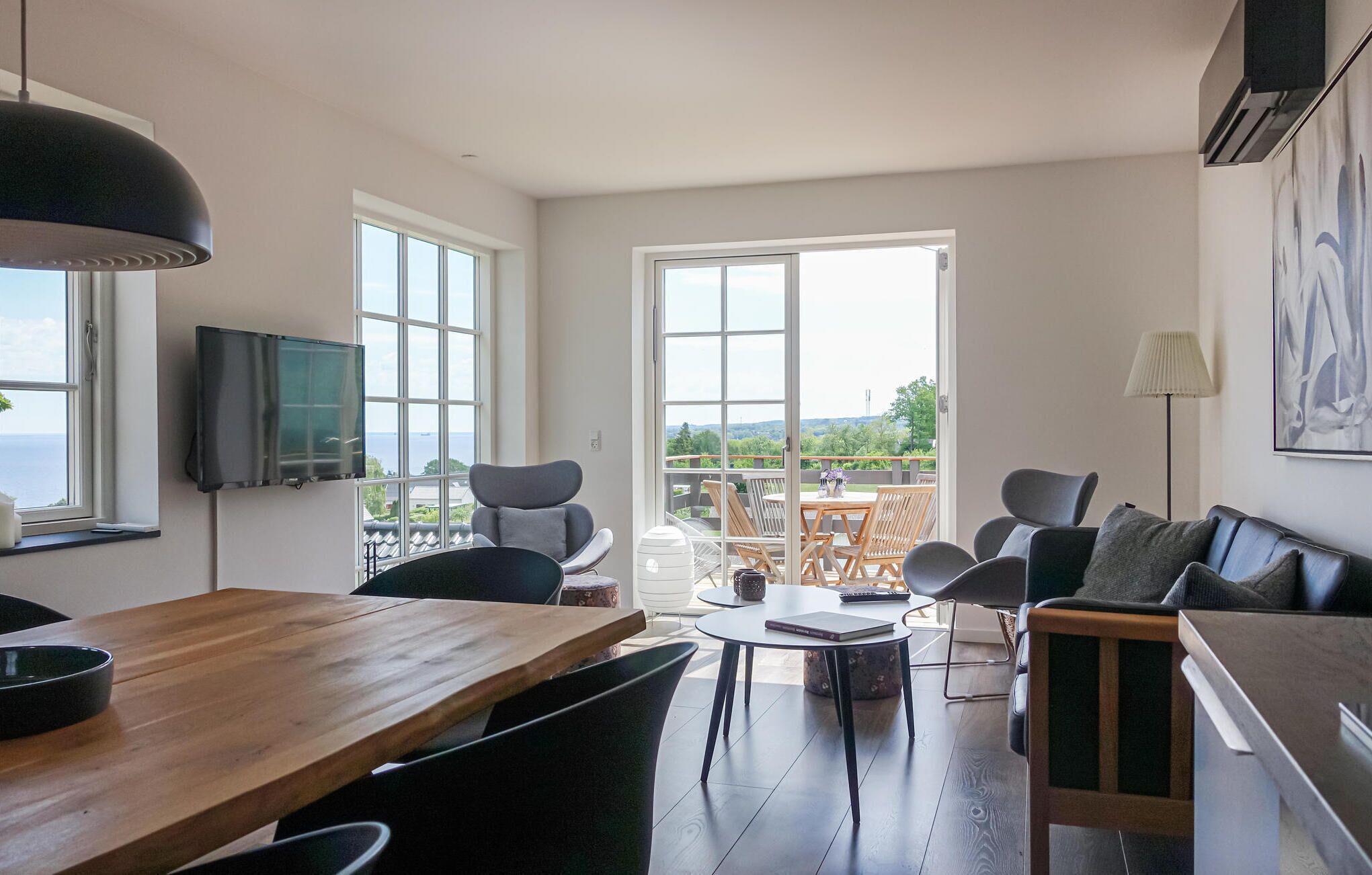 A modern living room with a wooden dining table, comfortable seating, a TV, and a balcony with a view of the sea.