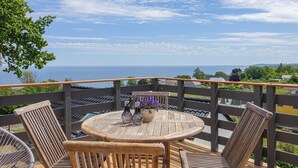 A wooden deck with a round table and two chairs, overlooking a scenic view of the sea and distant hills.