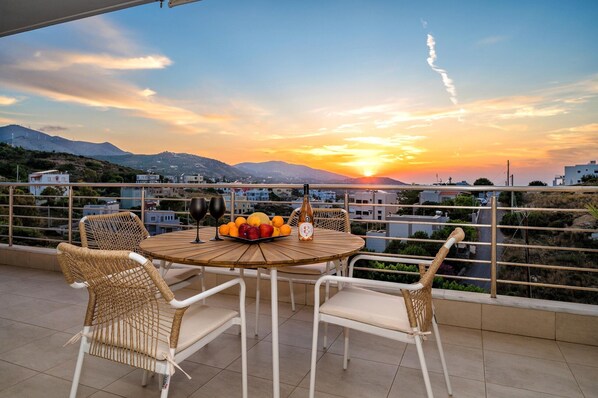 A round table with chairs on a balcony overlooking a sunset with mountains in the background.