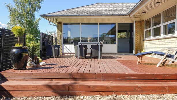 A wooden deck with a table and chairs, a large potted plant, and a view of the house through large windows.