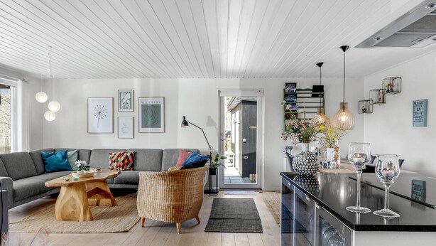 Una sala moderna con un sofá gris, una mesa de centro de madera y una silla de mimbre. Hay una zona de cocina con encimera negra y una copa de vino de cristal.