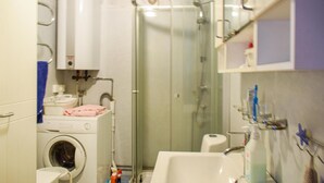 Une petite salle de bain équipée d’un lave-linge, d’une douche à parois de verre, d’un lavabo et d’une petite étagère où sont rangés les articles de toilette.