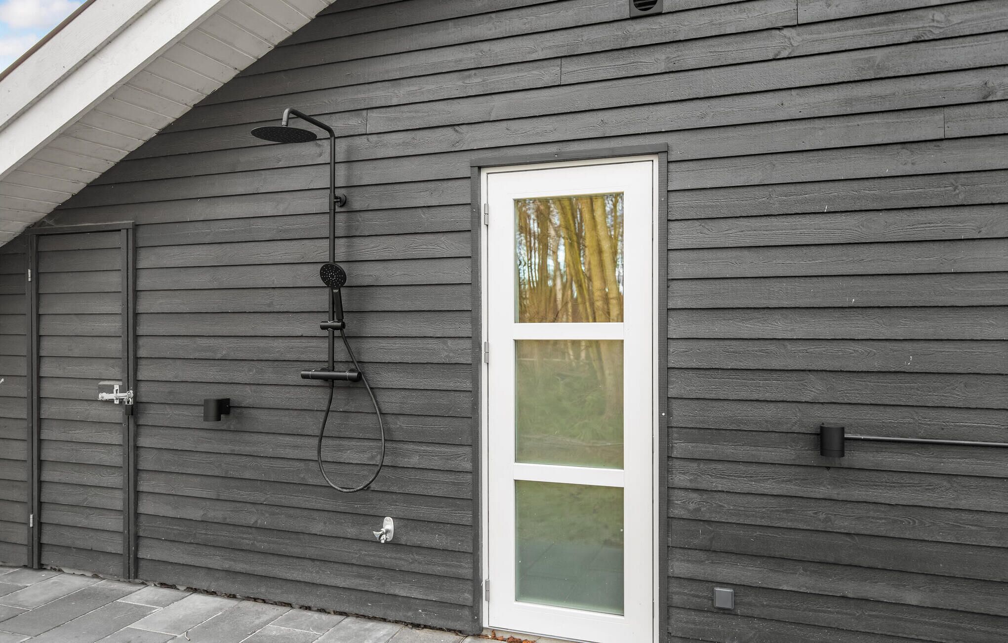 A gray wooden house with a white-framed door and a showerhead mounted on the wall.