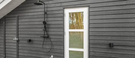 A gray wooden house with a white-framed door and a showerhead mounted on the wall.