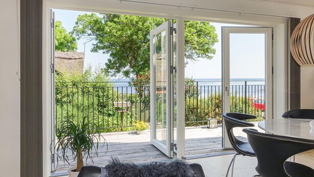 A balcony with a glass door, a table, and chairs, overlooking a body of water and greenery. - 2 bedroom cozy home in Svaneke (Svaneke)