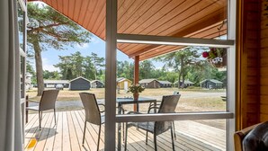 Um deck coberto ao ar livre com piso de madeira e cadeiras de vime, com vista para um camping com várias cabanas.