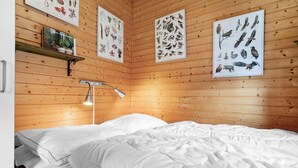 Quarto com paredes de madeira, uma cama com roupa de cama branca e três ilustrações emolduradas de plantas e pássaros na parede.