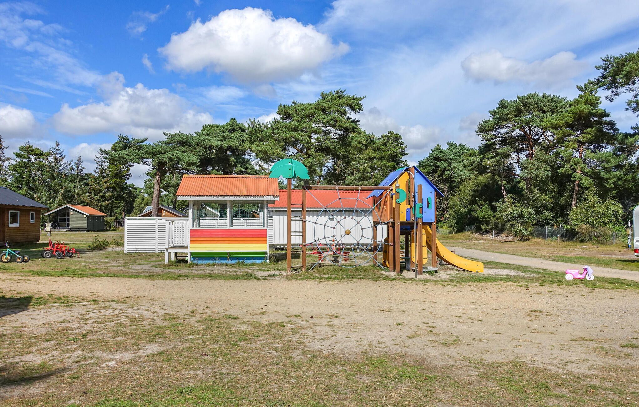 A playground with a colorful slide and climbing net, surrounded by trees and a small building with a red roof.