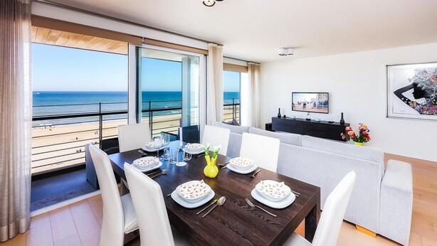 A dining area with a view of the sea, a sofa, a TV, and a painting on the wall. - Res. Vent de Sable ref.: 115 (Oostende)