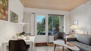 A modern living room with a white brick wall, a grey sofa, a dining table with chairs, and a large window with a view of greenery outside. - Amazing apartment in Allinge (Allinge)